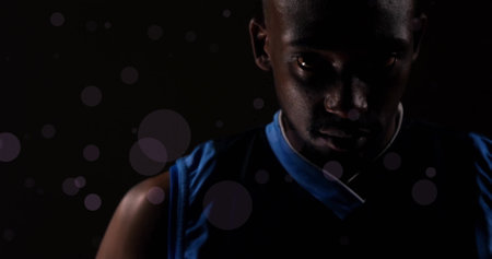 African American man wearing blue tank top, looking down. Sporting short black hair and dark eyes, standing in a dimly lit room with light spotsの写真素材