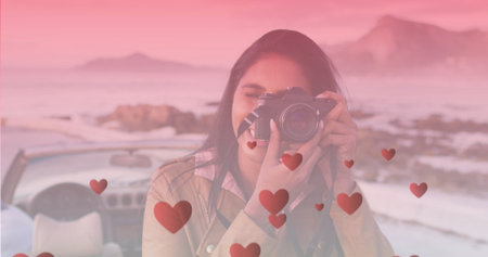 Image of hearts floating over happy biracial woman taking photo at cabriolet. travel, holidays,...