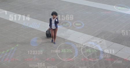 Image of numbers, circles, graphs over african american woman talking and walking with luggage. Digital composite, multiple exposure, business travel, report, progress and technology concept.の写真素材