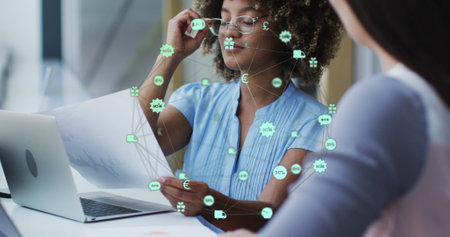 Image of globe of icons over two diverse female office colleagues discussing over a document. Business technology conceptの写真素材