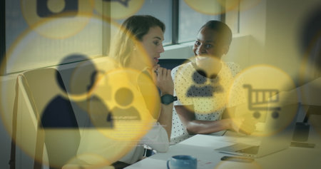 Image of multiple icons over diverse female coworkers discussing reports on laptop in office. Digital composite, multiple exposure, business, shopping, planning, teamwork and technology concept.の写真素材