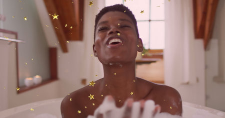 Image of gold stars falling over happy african american woman blowing foam in bath. wellbeing,...