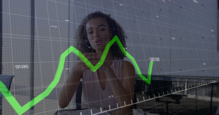 African American career woman wearing white tank top, standing by graph. With curly brown hair, examining financial data on a large screenの写真素材