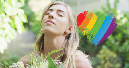 Image of rainbow heart over caucasian woman wearing wedding dress and holding flowers. Pride, lifestyle and celebration concept digitally generated image.の写真素材