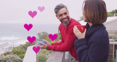 Image of hearts over diverse couple drinking coffe on balcony