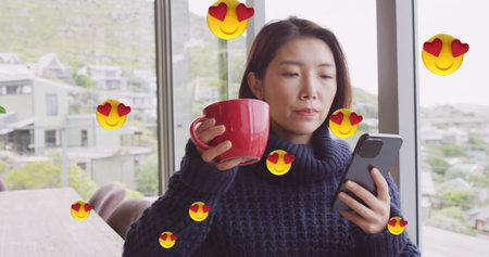 Image of emoji icons over asian woman drinking coffee and using smartphone at homeの写真素材