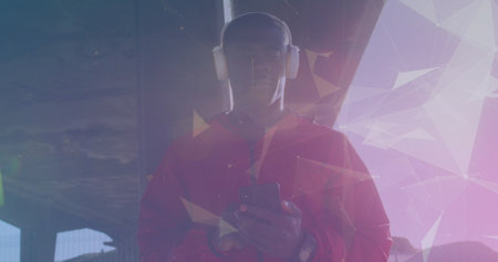African American male athlete wearing headphones, holding smartphone. Standing outdoors, looking...