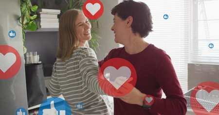 Image of hearts and like icons over caucasian female couple dancing. Valentines, love and...