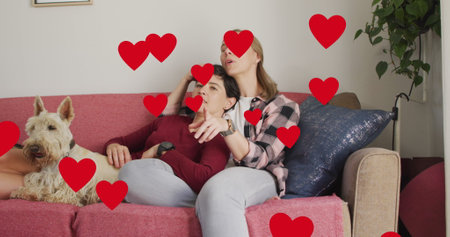 Image of hearts over caucasian female couple sitting on sofa with their pet dog. Valentines, love and celebration concept digitally generated image.の写真素材