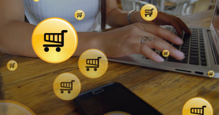 Image of shopping cart icon in circles, midsection of caucasian woman working on laptop. Digital composite, multiple exposure, retail, buying, shape, abstract and technology concept.の写真素材