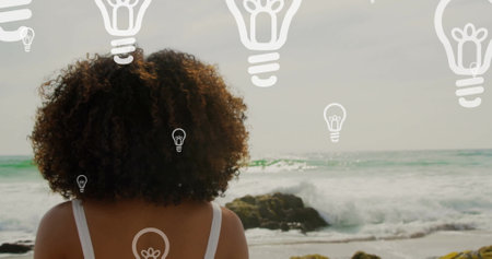 Image of light bulb icons over african american woman by seaside. Global social media, networks, digital interface and data processing concept digitally generated image.の写真素材