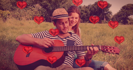 Image of red heart balloons floating over  man playing guitar for his woman in the field. Happy Valentines Day celebration concept digitally generated image.の写真素材