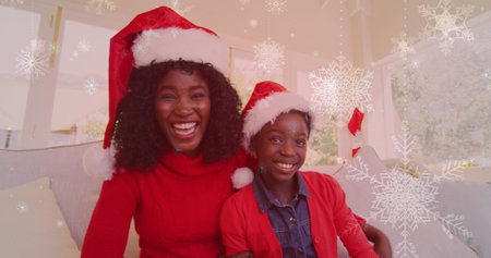 Image of snow falling over smiling mother and daugher with santa hats waving hands. christmas, winter, tradition and celebration concept digitally generated image.の写真素材