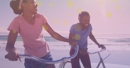 Yellow spots of light against african american couple sitting on their bicycles smiling at the beach. pedal day awareness conceptの写真素材