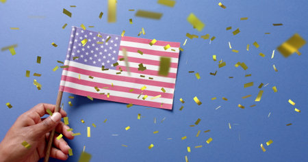 Image of confetti falling over hand holding flag of united states of america on blue background....