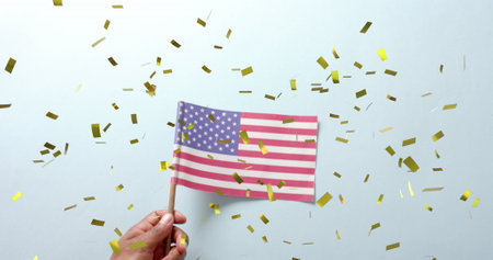 Image of confetti falling over hand holding flag of united states of america on blue background....