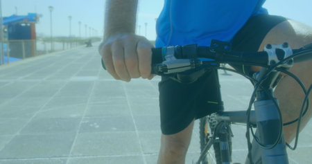 A senior Caucasian male is riding bicycle along a sunny promenade. He's wearing a blue shirt,...