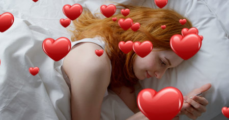 Digital composite of a Caucasian red haired woman lying in bed while texting and digital hearts flying in the foreground 4kの写真素材
