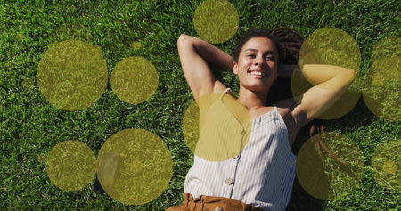 Image of yellow spots over smiling biracial woman lying in park. health and fitness concept digitally generated image.の写真素材