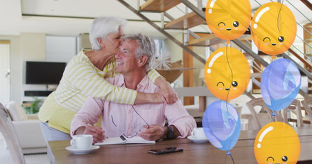 Image of party balloons over happy caucasian senior couple embracing in dining room at home. retirement lifestyle, celebration and wellbeing concept digitally generated image.の写真素材