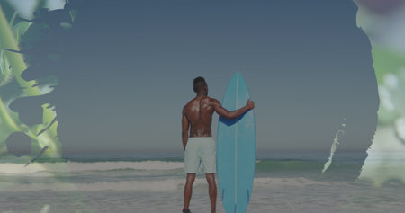 African American male holding surfboard on beach. He has dark skin, short hair, and is wearing white...