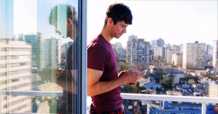 Man in purple shirt standing on balcony using smartphone, cityscape in background. Sunlight bathing...