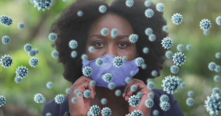 Image of coronavirus cells over african american woman putting on face mask in park. healthcare and safety during covid pandemic concept digitally generated image.の写真素材