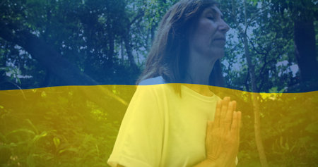 Image of flag of ukraine over caucasian woman praying outdoors. ukraine crisis and international politics concept digitally generated image.の写真素材