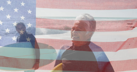 Image of flag of united states of america over senior biracial couple with surfboards on beach....