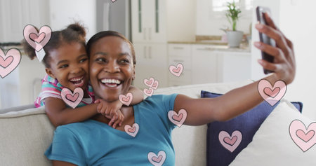 Image of happy african american mother and daughter taking selfie over hearts. family spending time...