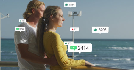 Image of social media icons over happy couple by seaside. global social media, connections, digital...