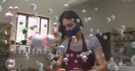 Image of virus cells over caucasian woman with face mask forming pottery. hobby during covid 19...
