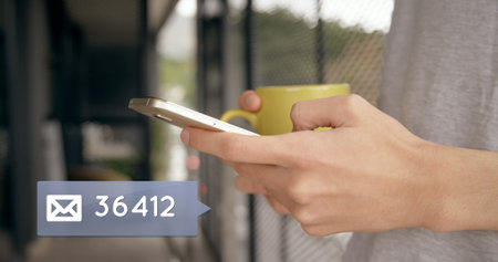 Close up of a hand typing on a mobile phone while the other hand is holding coffee mug. In the left...