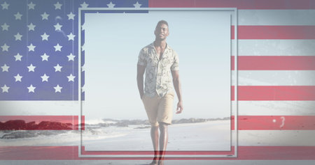 Young African American man walks on a beach with an American flag backdrop. He is wearing casual summer clothing and exudes a relaxed demeanor.の写真素材