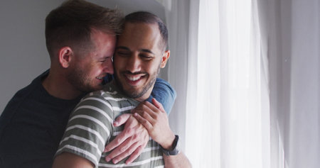 Diverse gay couple shares a tender moment, with. Embracing by the window, they enjoy a quiet, intimate connection at home.の写真素材
