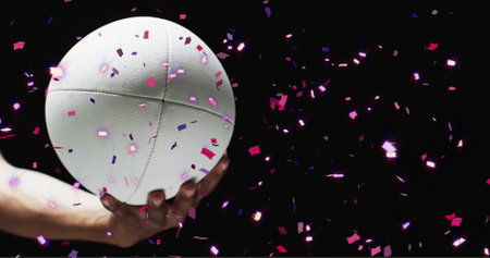 Image of confetti over white rugby ball held by caucasian man's hand on black background. Rugby, sports, competition and celebration concept digitally generated image.の写真素材