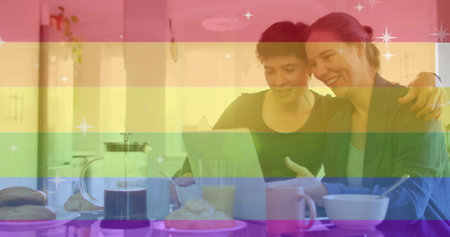 Image of pride text, rainbow stripes over happy caucasian lesbian couple using tablet at table. Domestic life, communication, lgbtq, gay, lesbian, pride and lifestyle, digitally generated image.の写真素材