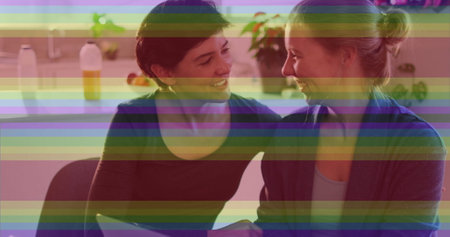 Image of pride rainbow stripes over happy caucasian lesbian couple using laptop at home. Domestic life, communication, lgbtq, gay, lesbian, pride and lifestyle, digitally generated image.の写真素材