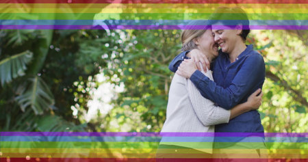 Image of pride rainbow stripes over happy caucasian lesbian couple embracing in garden. Romance, love, nature, lgbtq, gay, lesbian, pride and lifestyle, digitally generated image.の写真素材