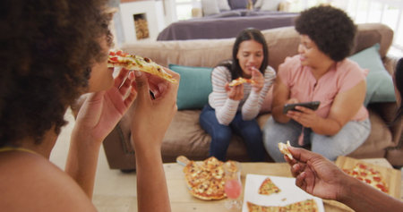 Happy diverse female friends sitting on sofa in living room, eating pizza. spending quality time at...