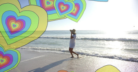 Image of colourful hearts over biracial woman walking on beach. Summer, vacation, lifestyle and...