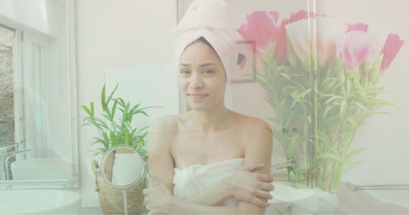 Image of pink flowers in vase over portrait of happy biracial woman wearing towel in bathroom....