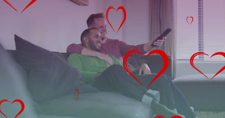 Image of heart icons over diverse gay couple smiling and watching tv. pride month and celebration concept digitally generated image.の写真素材