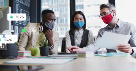 Image of social media icons over diverse colleagues wearing face masks discussing at office. Social media networking and covid-19 pandemic conceptの写真素材