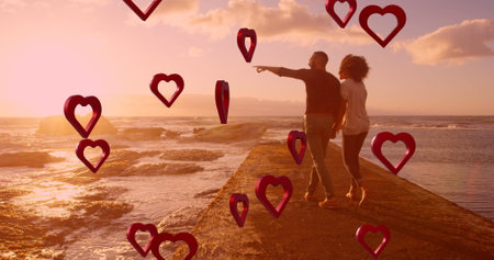 Image of red hearts over couple in love holding hands by sea. Valentine's day, love and romance concept digitally generated image.の写真素材