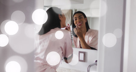 Image of light spots over biracial woman applying lipstick. Lifestyle, fashion and light concept digitally generated image.の写真素材