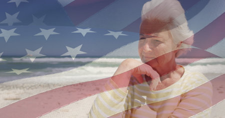 Image of senior caucasian woman sitting on beach over flag of united states of america. american patriotism, holiday and retirement concept digitally generated image.の写真素材