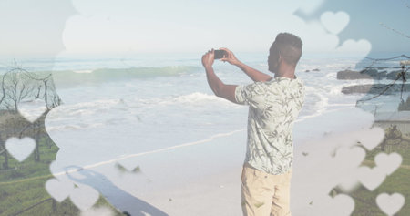 African American male capturing beach scenery with smartphone. He's wearing patterned shirt, khaki shorts, and has short hairの写真素材