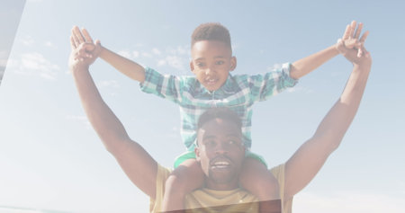 African American father lifting son on shoulders, both smiling under clear sky. Father wearing...