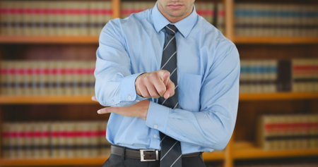 Caucasian male, wearing blue shirt and tie, checking time in library. Short brown hair, sporting a wristwatch, surrounded by booksの写真素材